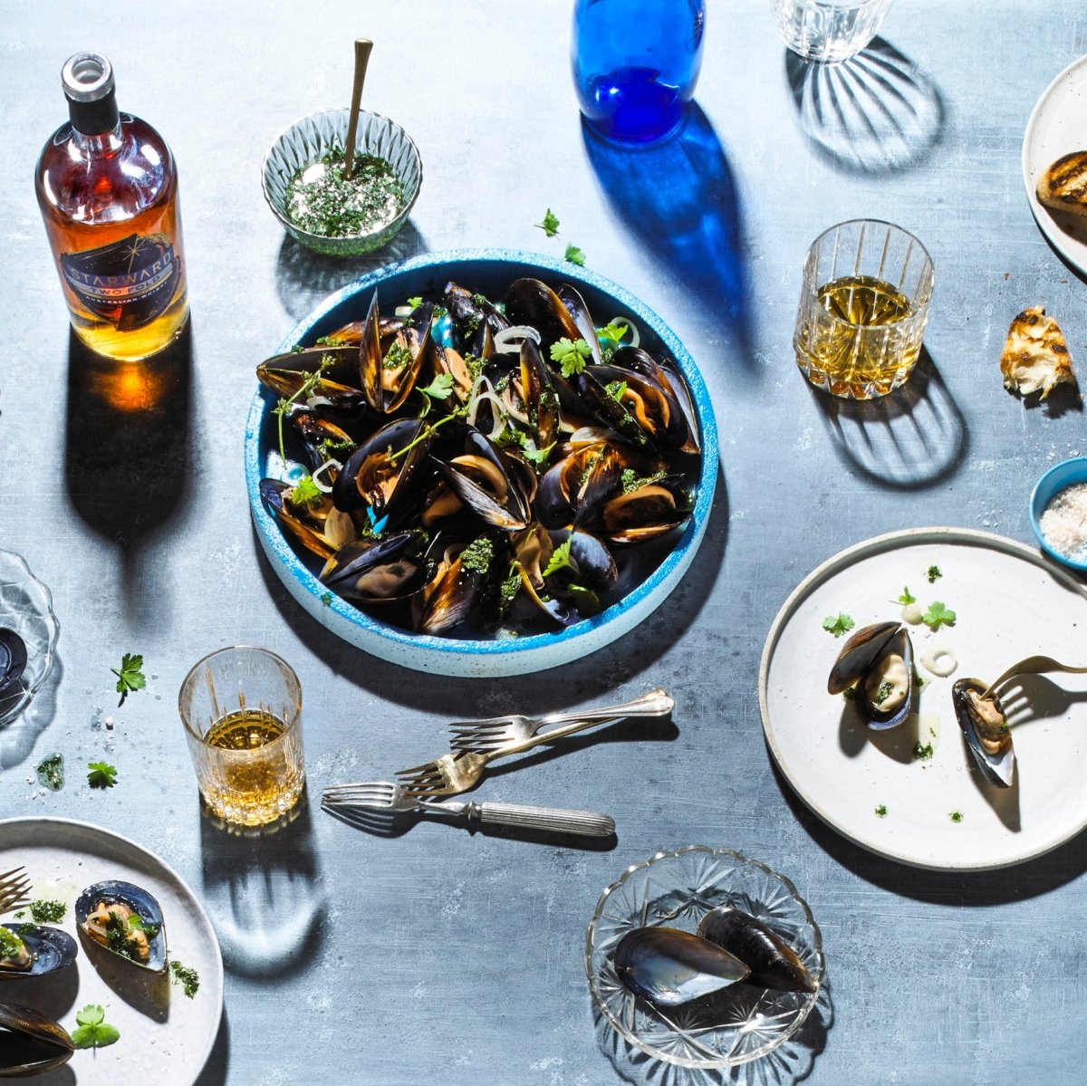 dinenr spread food blue glass whisky bottle liquids gold amber bright light paltes forks knives mussels seafood black shells