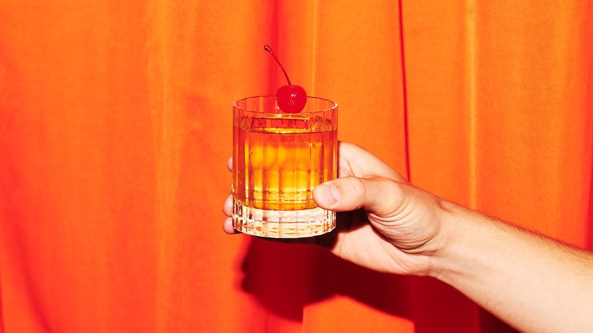 hand holding whisky glass cocktail red cherry garnish orange bright curtain