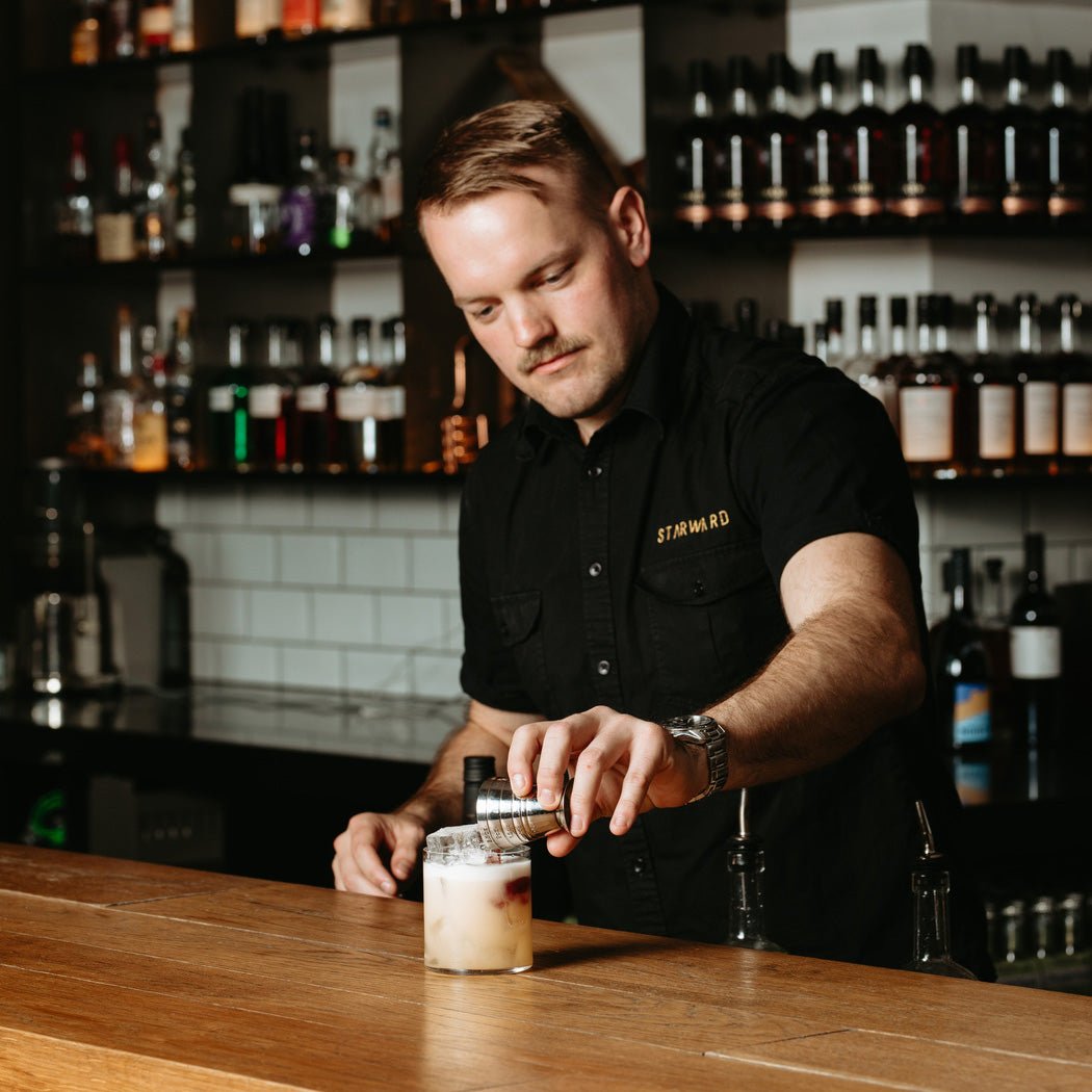 man bar pouring liquid cocktail layers red wine whisky yellow wooden bar shelves whisky bottles