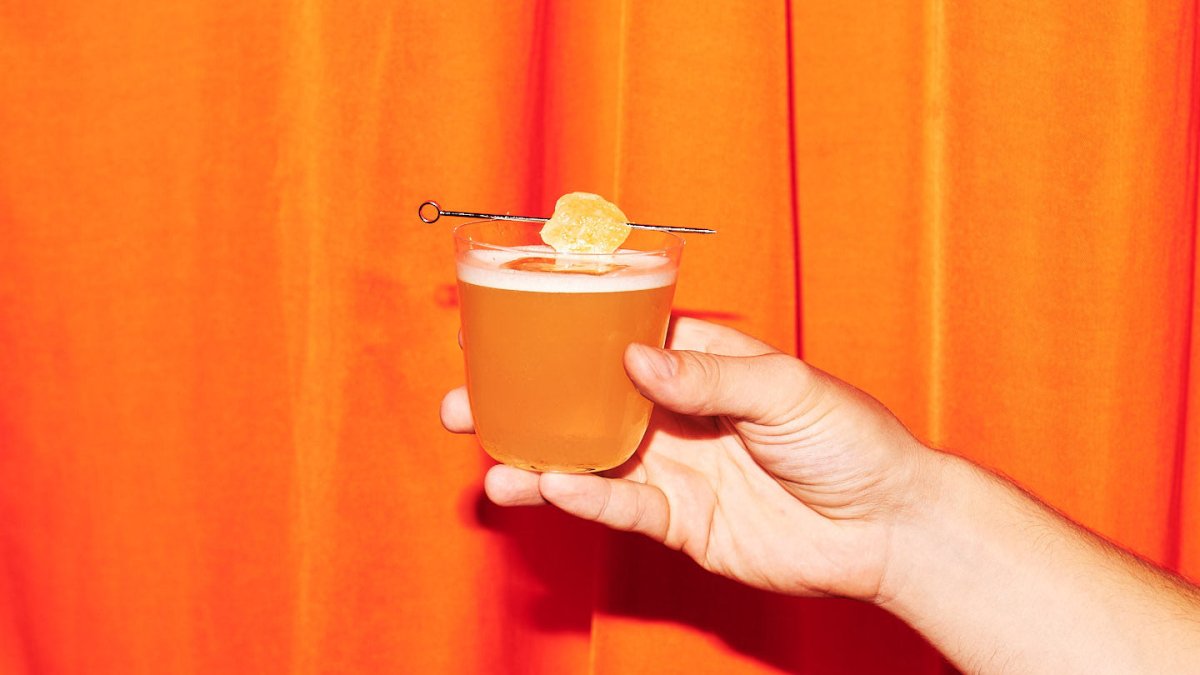 hand holding whisky glass cocktail skewered candied ginger garnish orange bright curtain