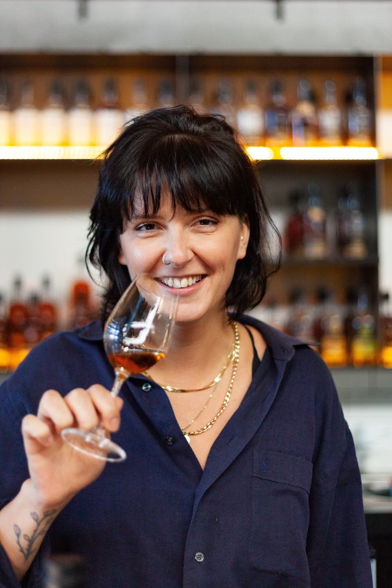 smiling woman blue shirt holding whisky glass amber liquid shelf whiskies spirits